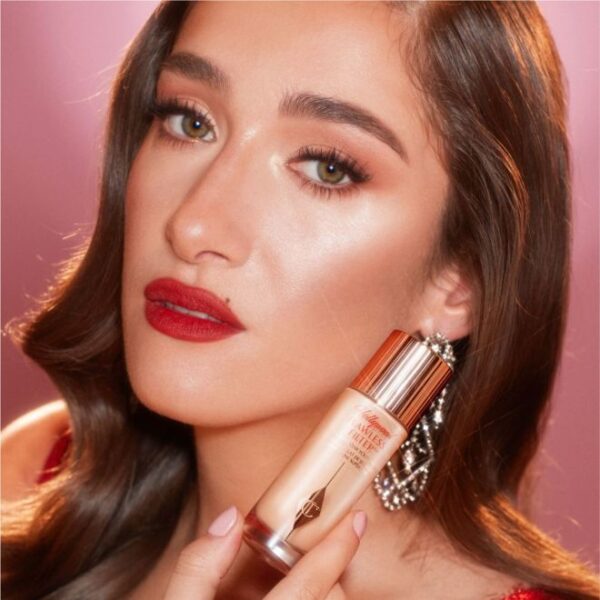 Vrouw met rode lippen en glamoureuze make-up houdt een fles foundation vast tegen een roze achtergrond.