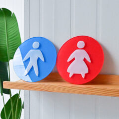 Pictogrammen voor toilet: blauw voor mannen, rood voor vrouwen, op houten plank naast groene plant.