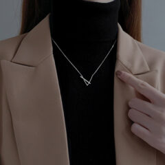 Minimalistische zilveren ketting over zwarte coltrui en beige jas, vrouwelijke hand wijst ernaar.