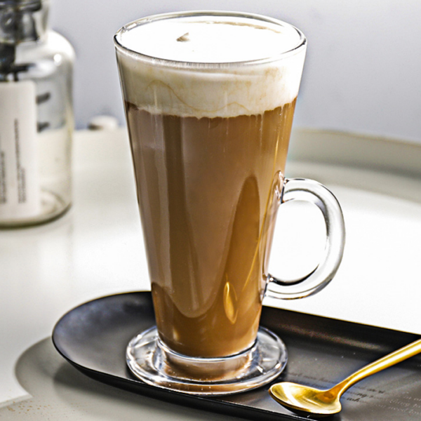 Lange latte macchiato in een glazen mok met melkschuim, geserveerd op een schotel met een gouden lepel ernaast.