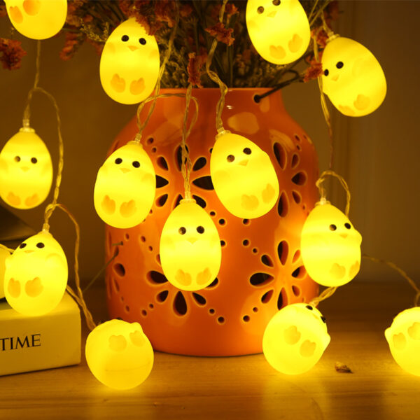 Gele kuiken-slingerlichten hangen aan een oranje vaas voor gezellige paasdecoratie in een warme huiskamer.