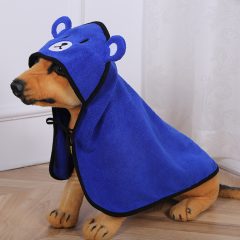 Hond in blauwe berenkostuumcape zit op houten vloer bij gordijn.
